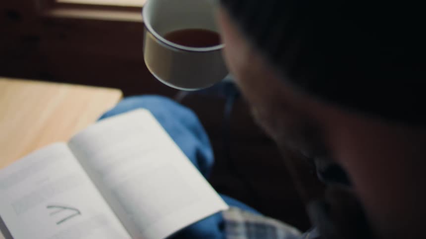 Man sitting near window reads a book while drinking from a mug