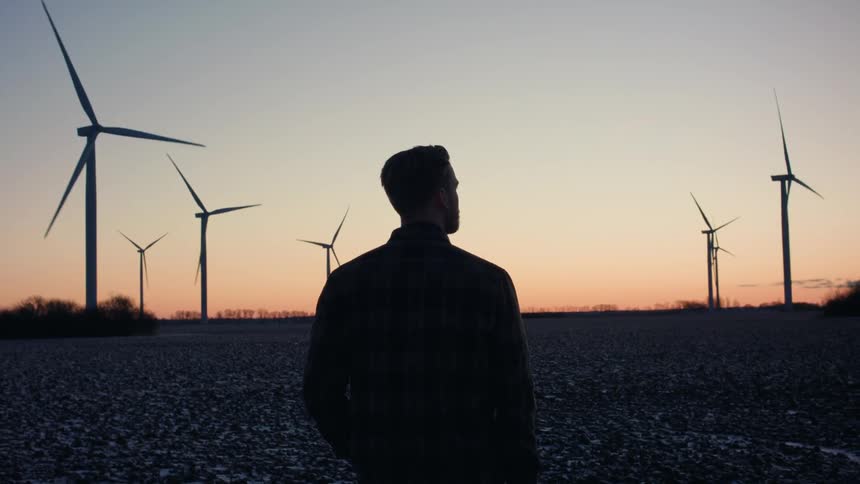Man stands looking at wind turbines partially silhouetted against a red horizon during winter