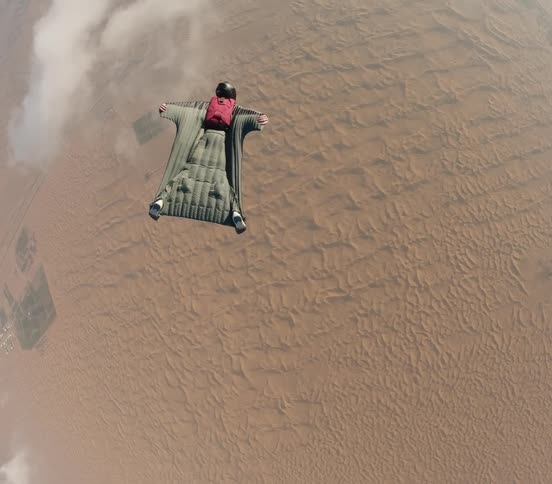 Person wearing wingsuit glides among clouds over arid terrain