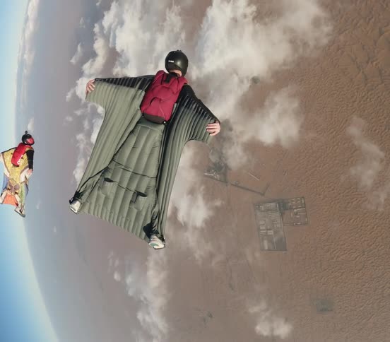 Two people wearing wingsuits glide over desert on a cloudy day