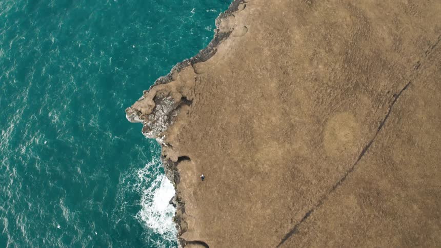 A person walking next to a cliff that falls straight into the sea and there are several birds flying over the place