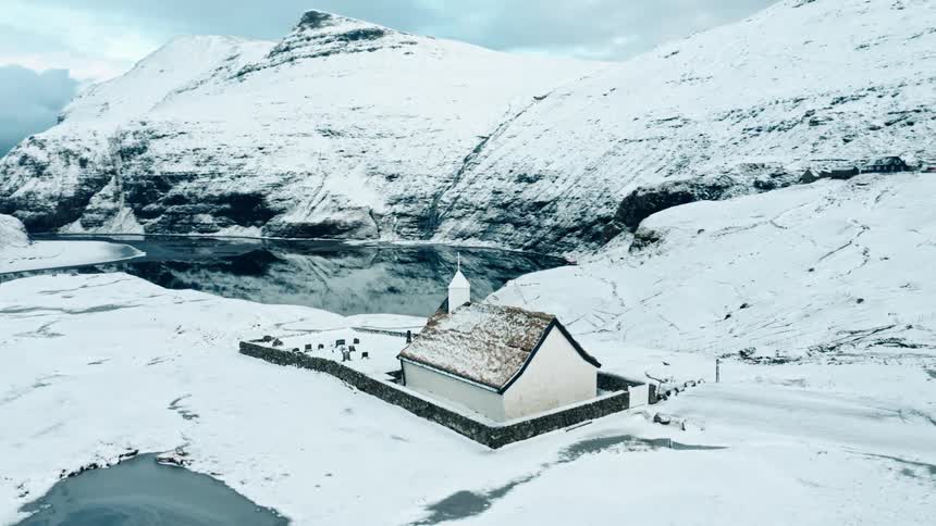 Church with snow covered graveyard sits on hill across pond from mountains during winter