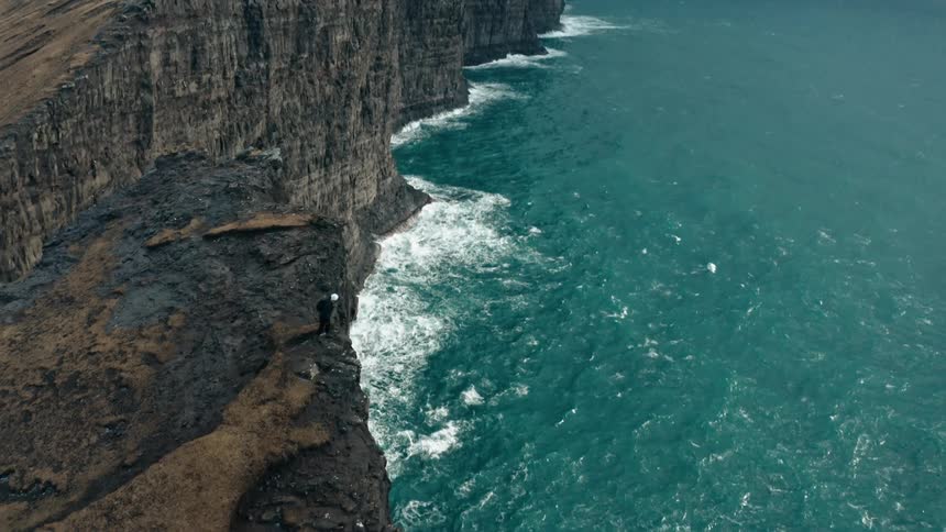 Aerial view of a person on the cliff of a promontory observing a turbulent sea on a cloudy day