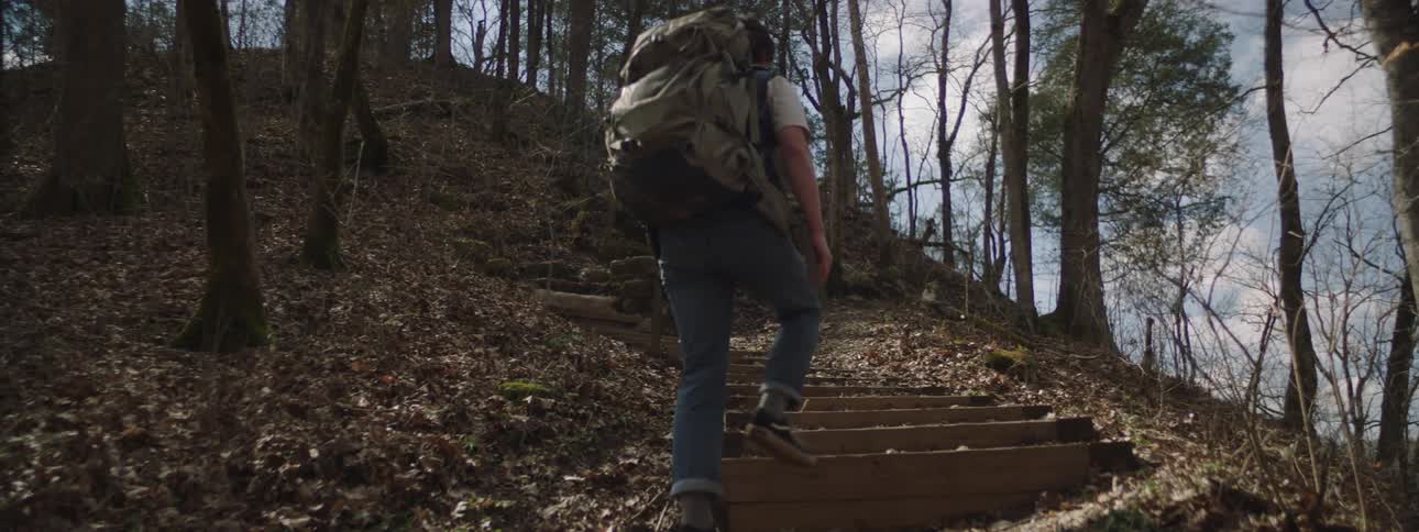 A man with a huge backpack is climbing a staircase and there are many trees around him