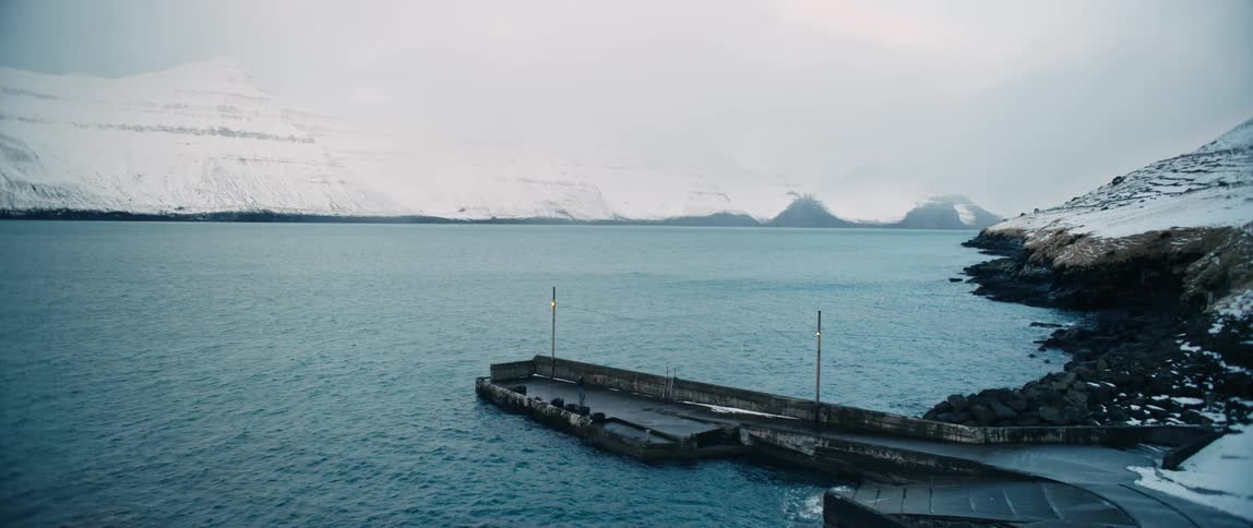 A person walks on the dock along a wide river with snow covered mountains in the background