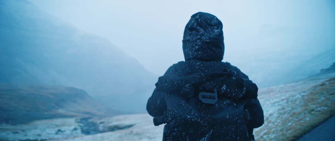 A person wearing a jacket is on an asphalt street looking at the horizon and it is snowing