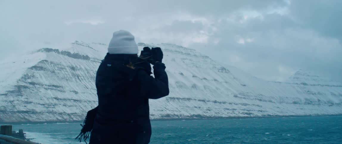 An adult takes a photo of the ocean with snow covered mountains against the shore