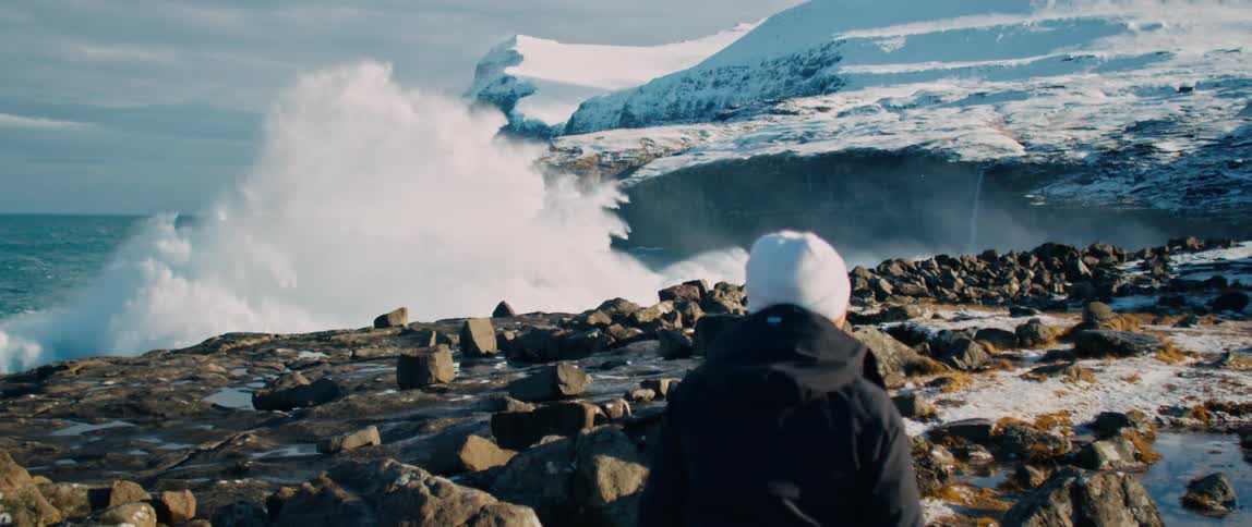 A person will have their back turned and look at the waves crashing on a coast full of rocks