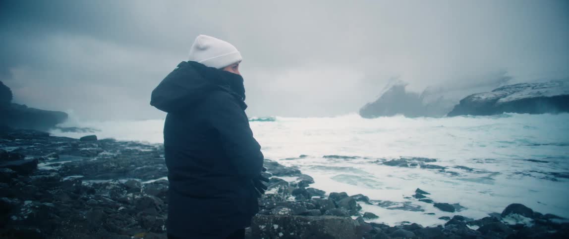 A person in a cold coat is looking at the sea coast with several waves breaking on the rocks