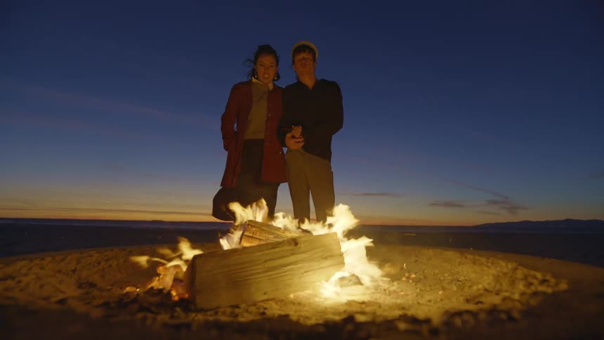 Two friends looking at a campfire in the beach sand at evening