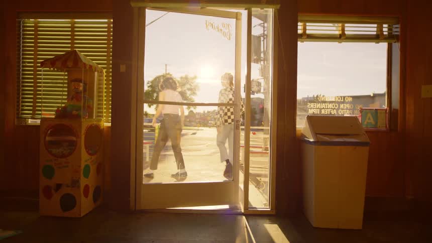 Two people are walking up to a door and leaving an establishment and there is traffic and bright sunlight outside