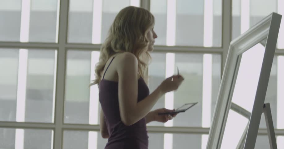 A woman stands at a mirror applying makeup