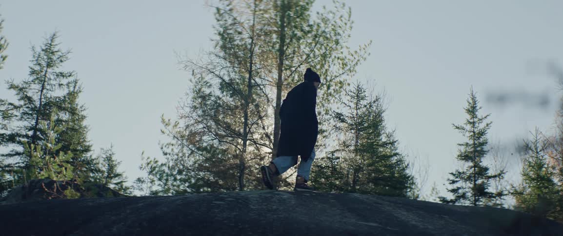 A woman walks through the sunshine on top of a large flat rock by a forest