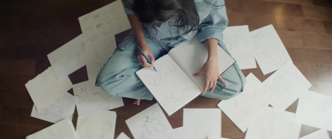 Female with dark hair wearing denim clothes sits on floor drawing in notebook