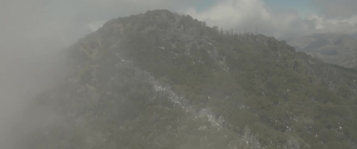 Aerial flyover rock mountain peak with vegetation partially covered in fog during the day