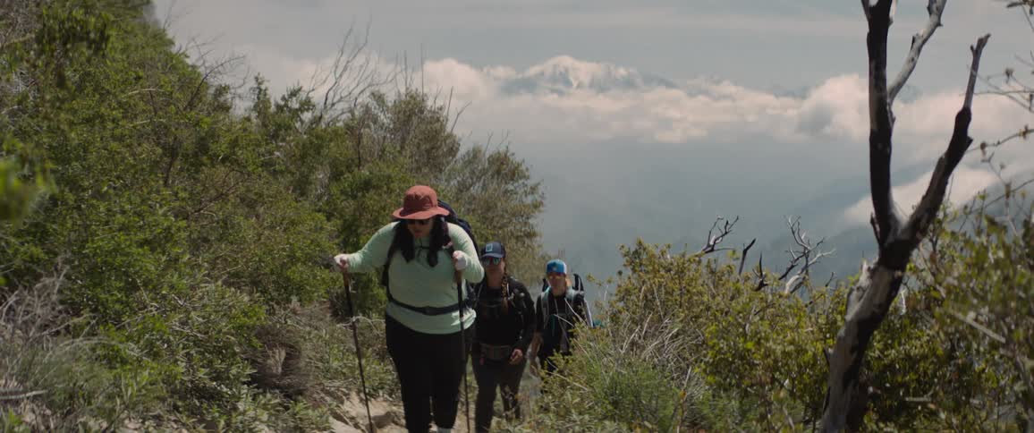Hikers walk along trail on mountain across valley from snow covered mountain on a cloudy day