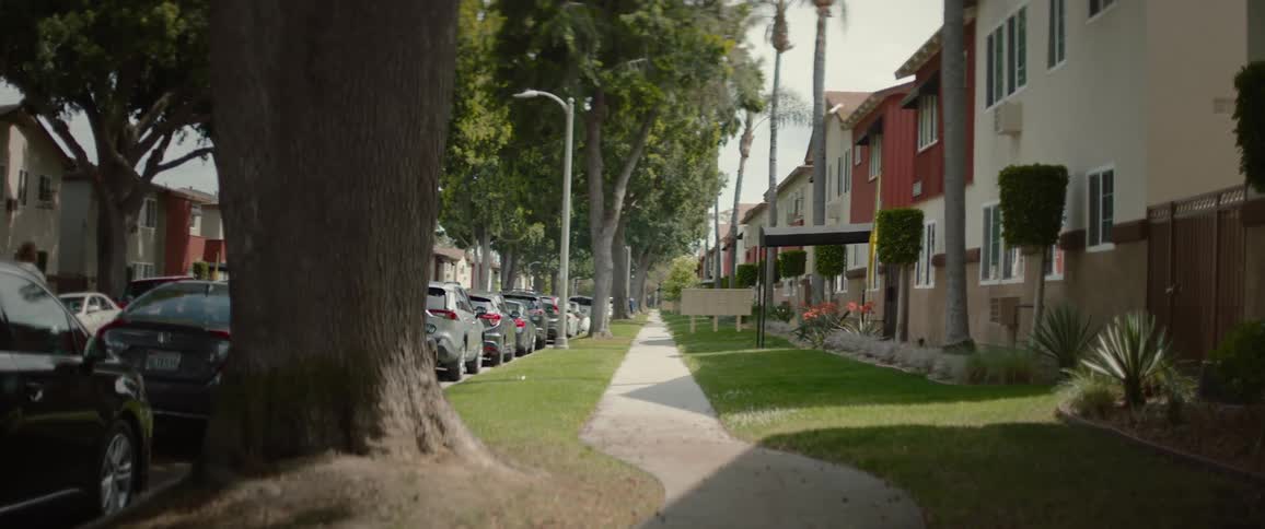 A sidewalk lined on one side with closely built town home apartment buildings and a man running down the street lined with cars on the other side on a sunny afternoon