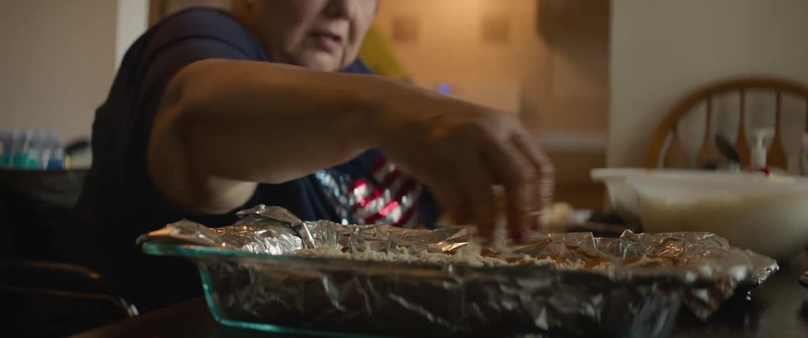 Woman sitting at a dinning room table sprinkling cheeses into a nine by eleven dish in natural sunlight as another woman walks back and forth in the kitchen in the background