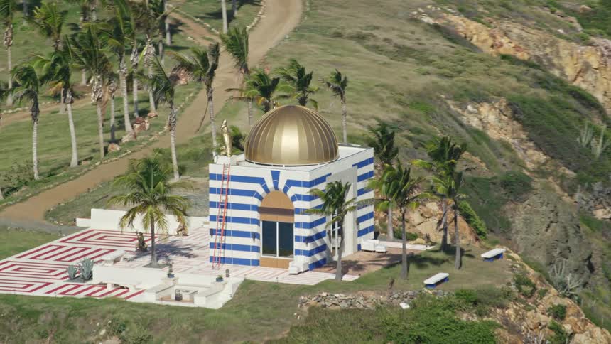 A white building with a bronze dome and geometric designs in blue surrounded by palm trees on the edge of a cliff by the sea