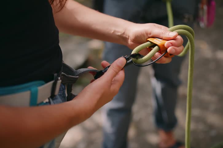 Woman standing up outdoors adjusting hooks on a hiking rope next to another standing person during day time