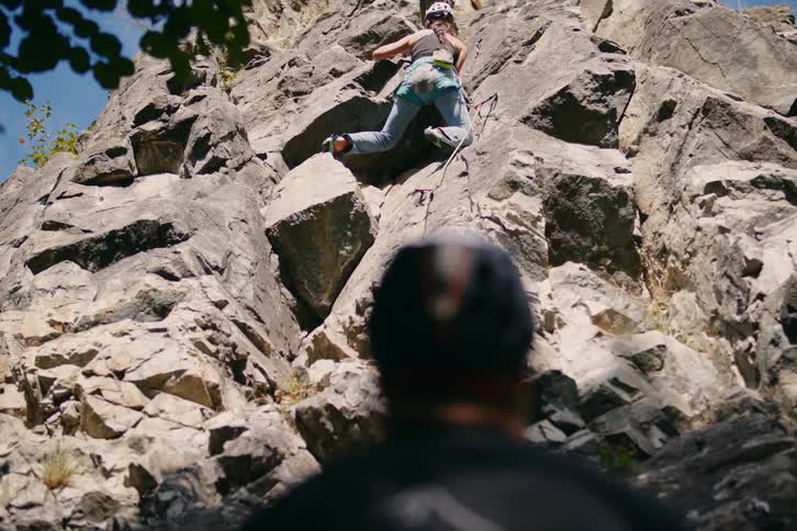 View of a mans back watching a woman climb a rocky wall of a mountain