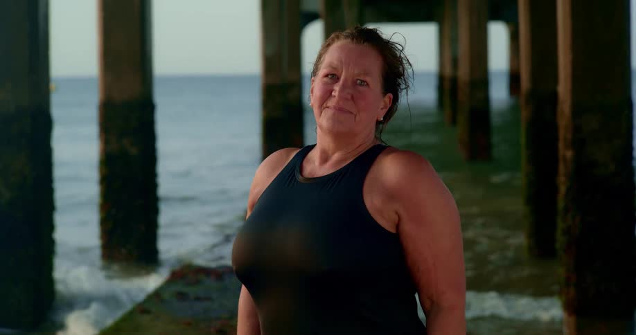 Mature woman wearing a black swimsuit under a pier next to the ocean