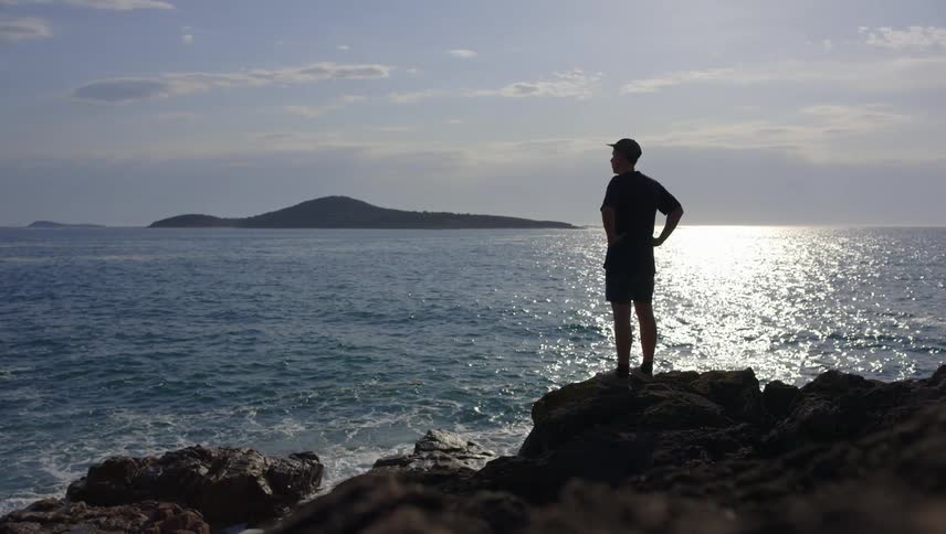 A man standing on a rock close to shore in the evening