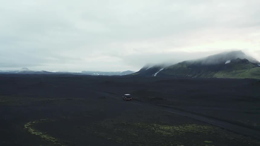 Vehicle traveling through natural black terrain with a chain of mountains around on a cloudy winter day