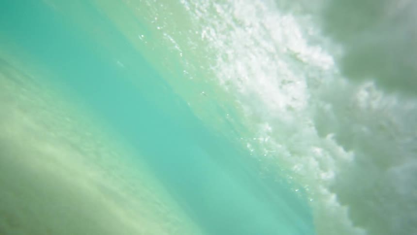 A large wave of clear ocean water crashes down upon the sandy beach