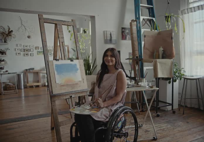 Female artist sitting in wheelchair looks up to smile while painting in art studio