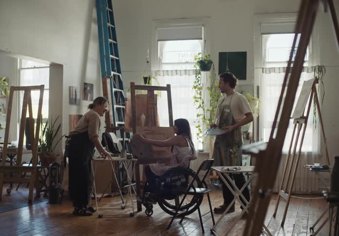 Three people in a room with one of them a woman in a wheelchair talking about painting