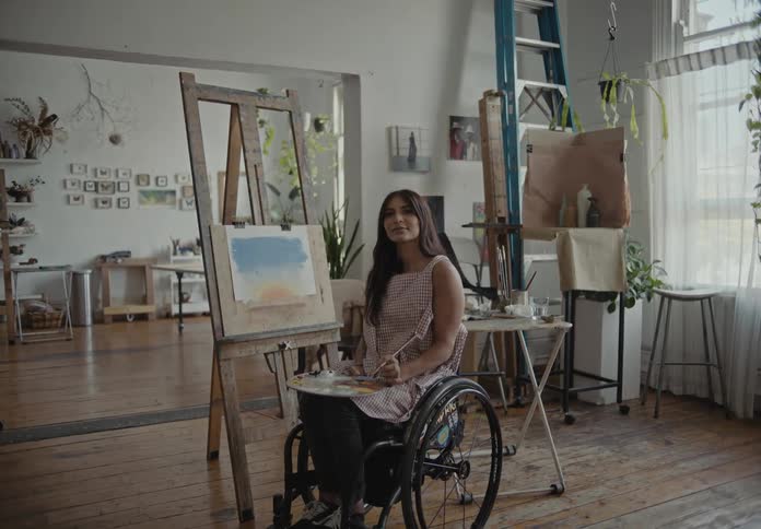 Female artist sitting in wheelchair smiles while holding palette and paintbrush in art studio