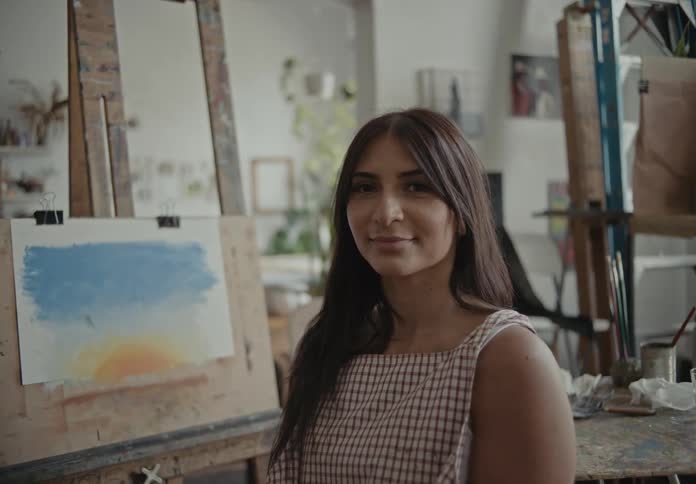 A woman sitting down and preparing to continue painting gives the camera a smile