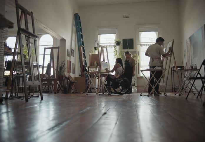 Wheelchairbound woman and another painter in an indoor room painting pictures with a woman supervising