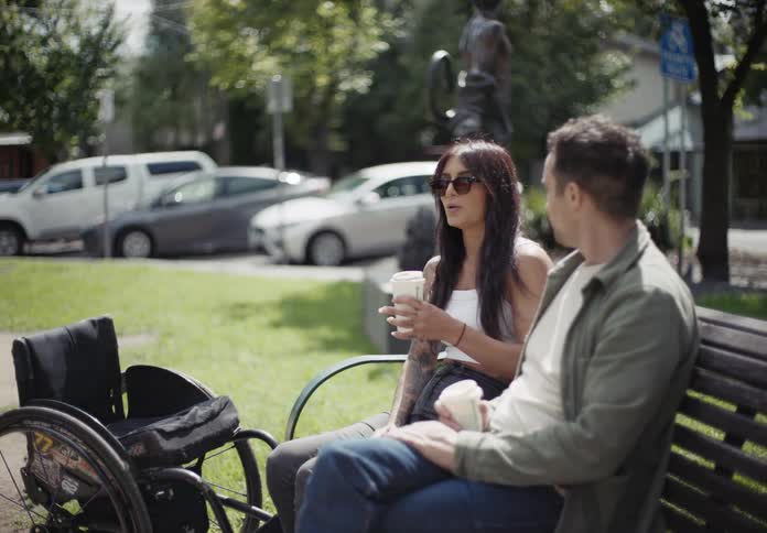 Two friends holding disposable coffee cups sit on park bench near empty wheelchair and talk