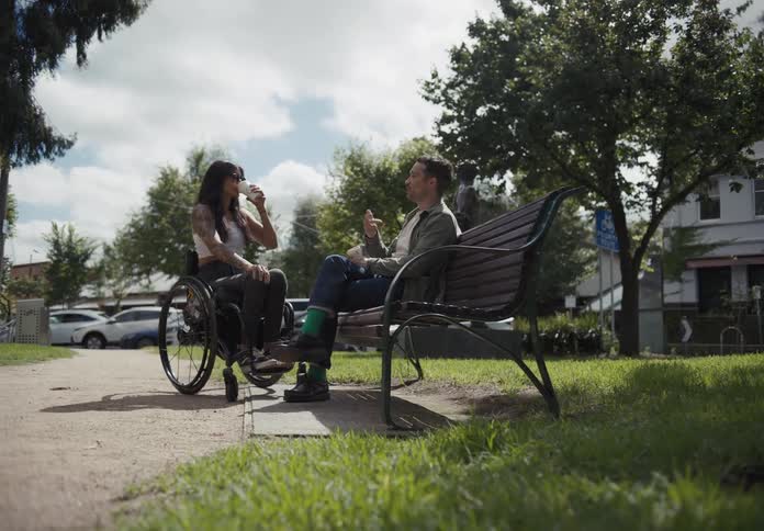 Man sits on park bench talking to woman sitting in wheelchair as she sips from disposable cup