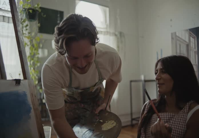 A standing man is painting a picture and playing with a woman who is in a wheelchair and they both have fun passing the brush over their faces