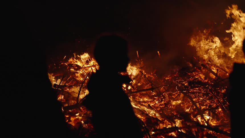 The silhouette of two people in front of a burning of several sticks with a very high fire