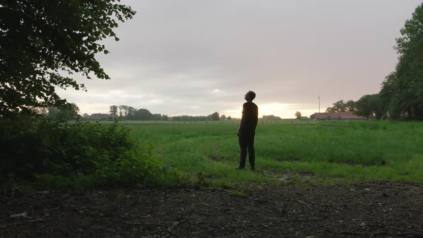 A man stands in a rural field admiring the sky at sunset