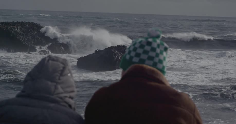 Two people sit bundled up on the shore as waves crash into rocks