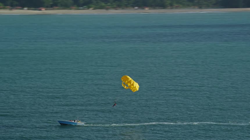 A boat in a sea with a rope tied to a person who is parachuting yellow behind the boat