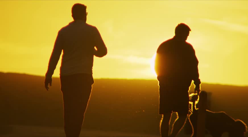 Men silhouetted against setting sun walk a dog