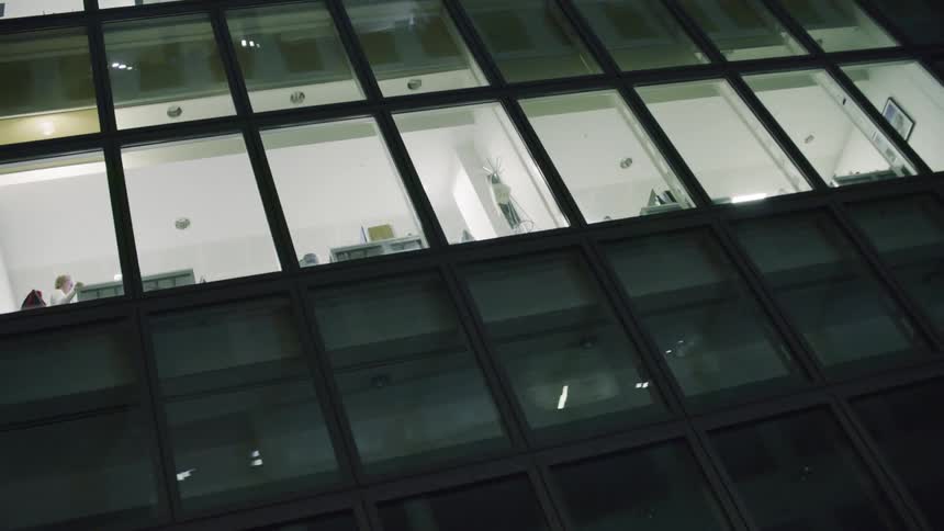 Female employee stands in brightly lit office in building with reflective windows at night