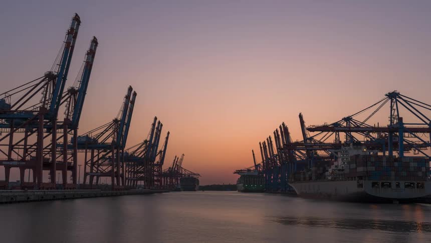 Loading cranes work over container ship as multiple watercrafts leave channel while night falls
