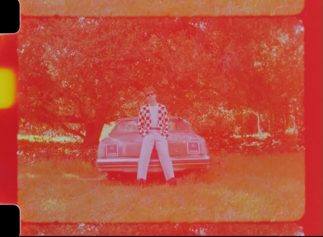 A young white male leans on a car near a forest
