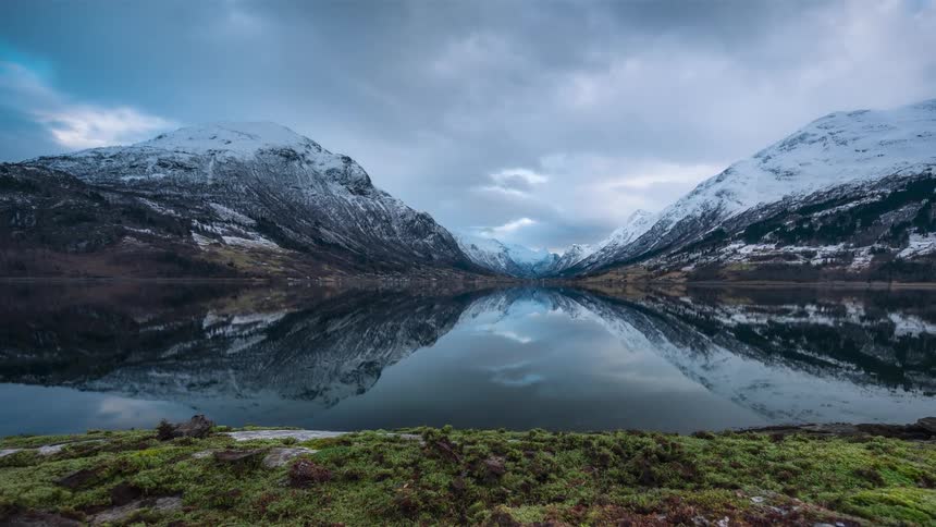 A serene scene of a calm lake mirroring the cloudy sky and the snowcapped mountains