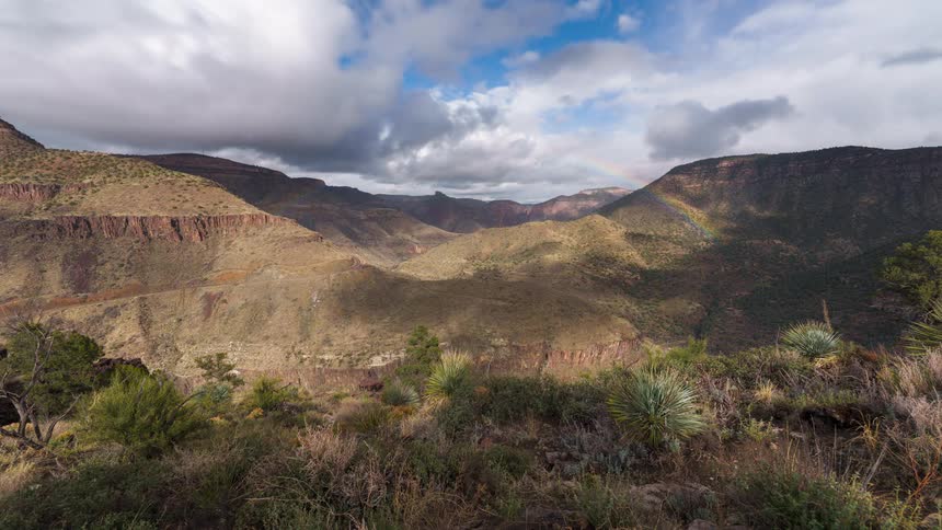 A mountain range with a subtle rainbow and green vegetation under a patchy sky