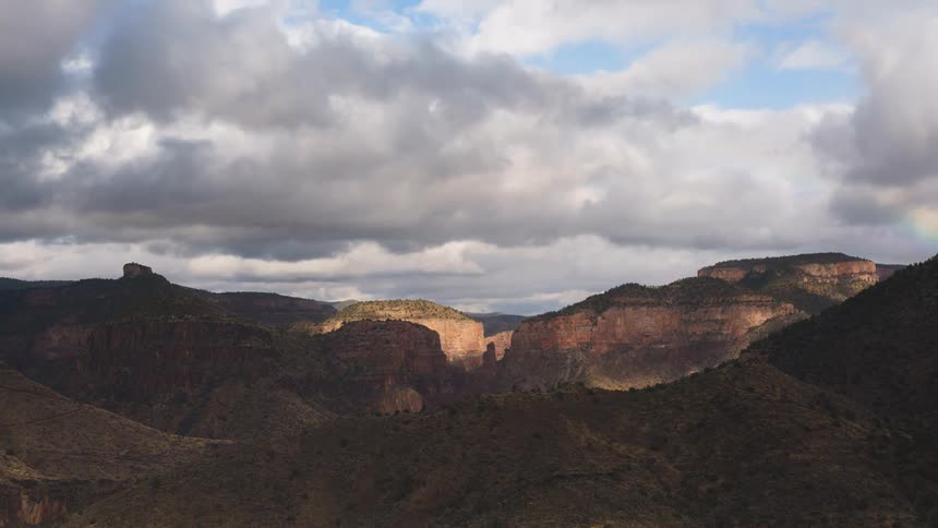 Rainbow curves over mountains as clouds pass overhead casting shadows onto the terrain