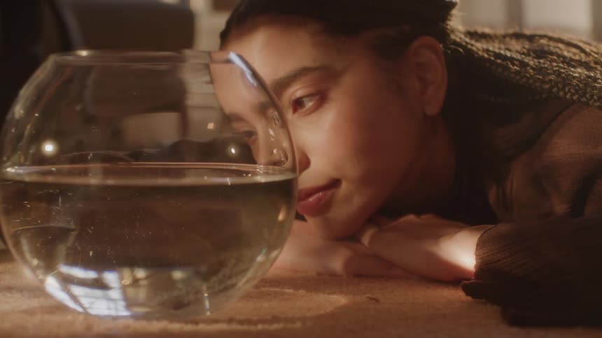 A young woman lays on the floor watching a goldfish swim around a bowl