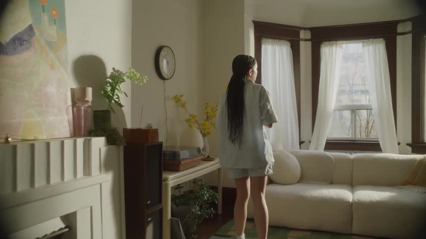 A young woman in pajamas walks into the living room with a cup of coffee